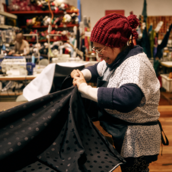 A woman with red beanie and glasses sews a dark polka-dot garment by hand in a cluttered workshop filled with sewing equipment.